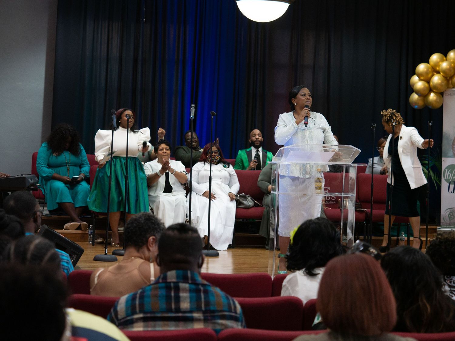 Church choir members in white and teal robes perform during a service with gold and white balloon decorations.