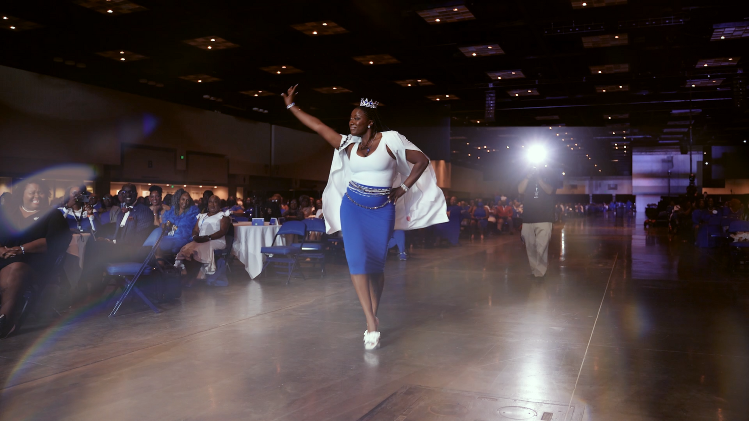 A dancer performing an energetic move on a dimly lit stage with dramatic lighting and audience in background.