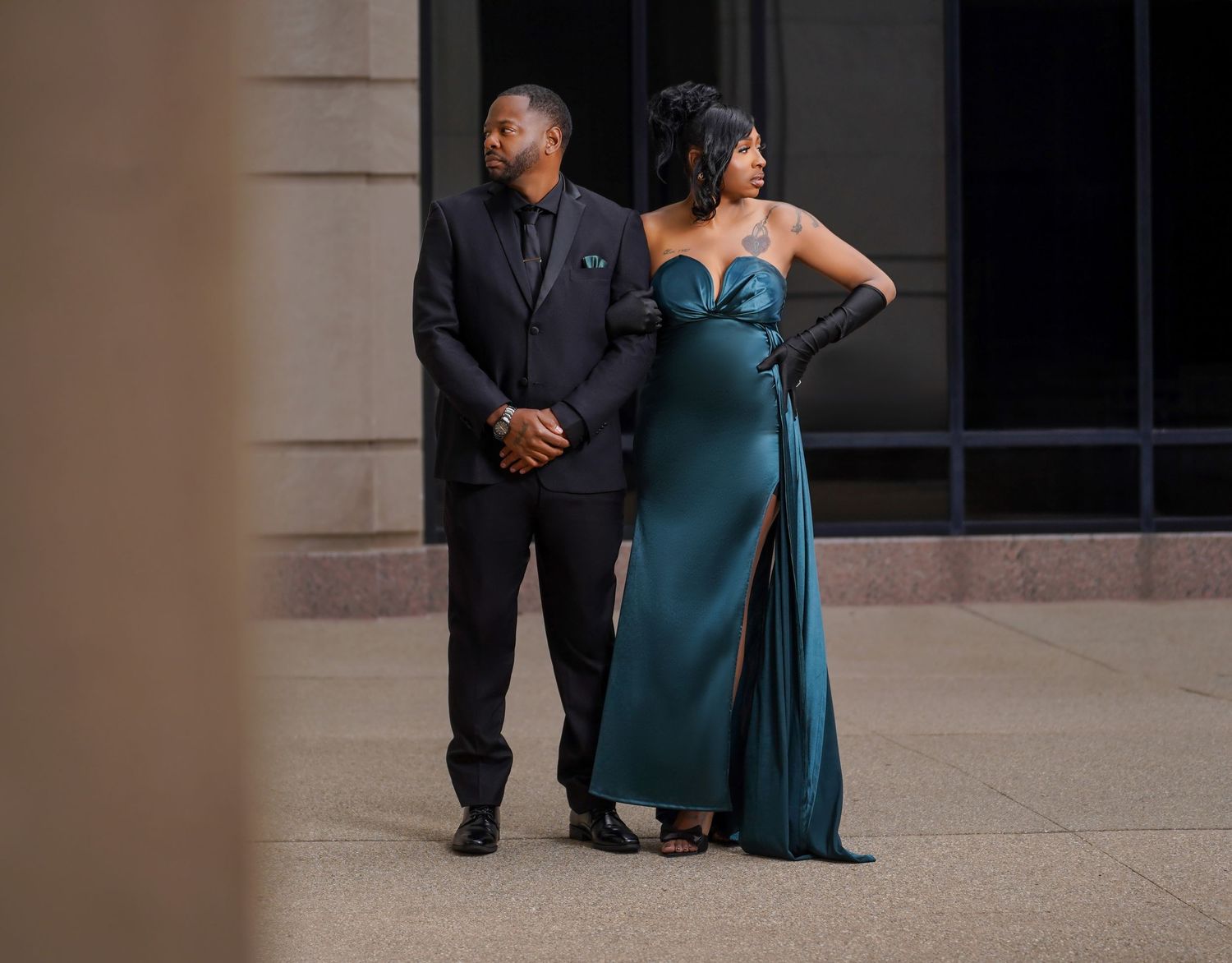 A formal couple poses outdoors, with someone wearing a black suit and another in an elegant teal evening gown.