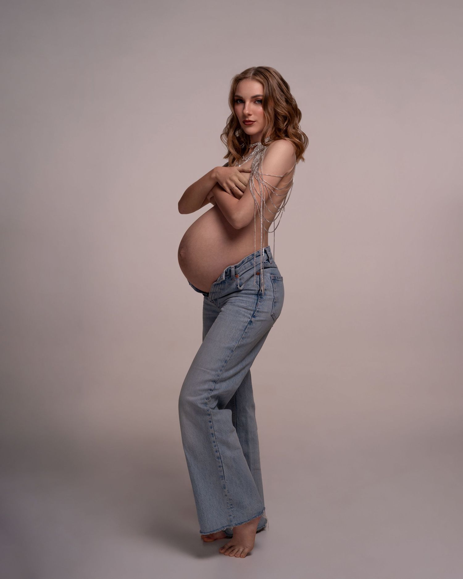 Topless pregnant women in jeans and crystal necklace posing holding hands over the breast.