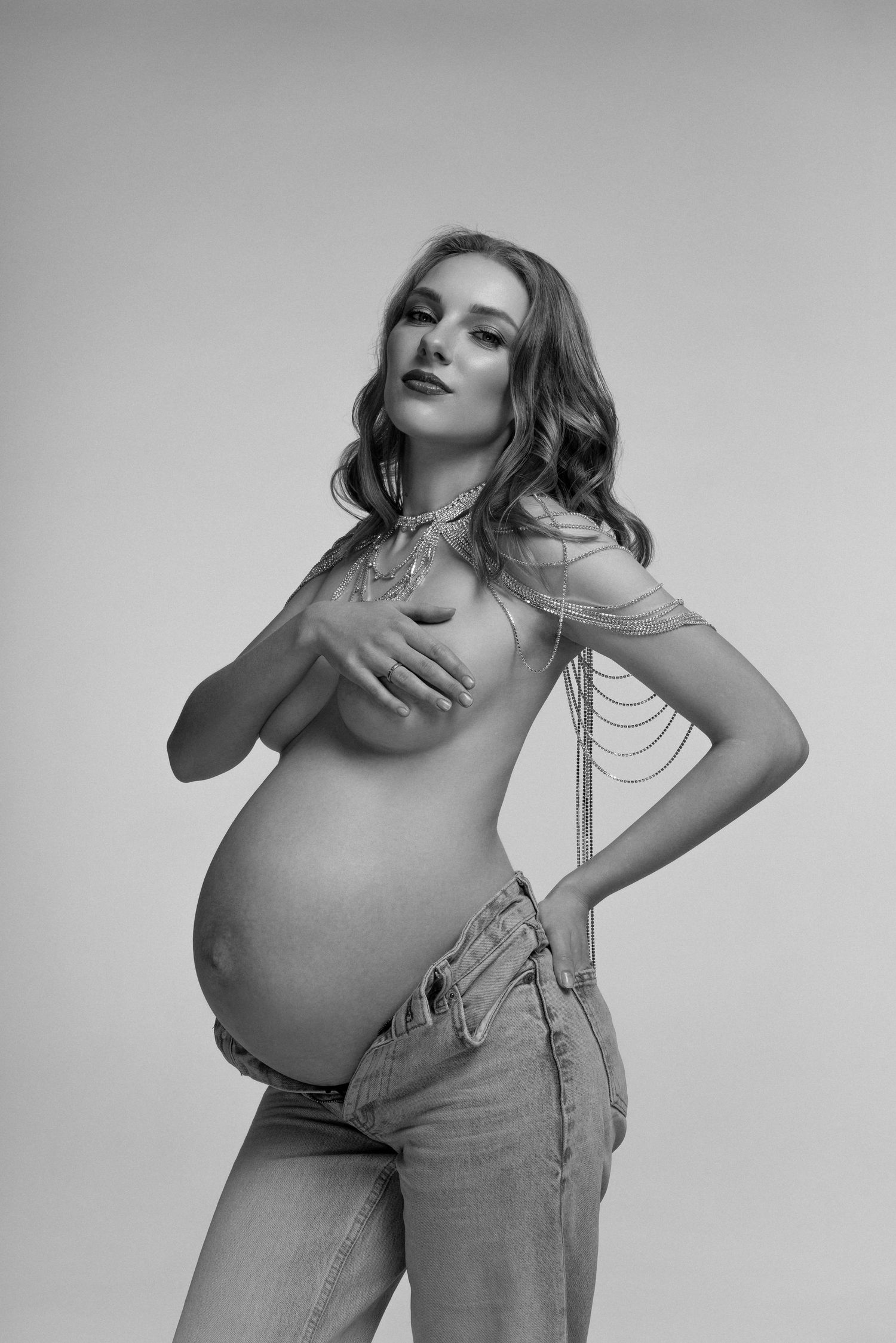 Black and white shot of topless pregnant women in jeans and crystal necklace posing holding hands over the breast.