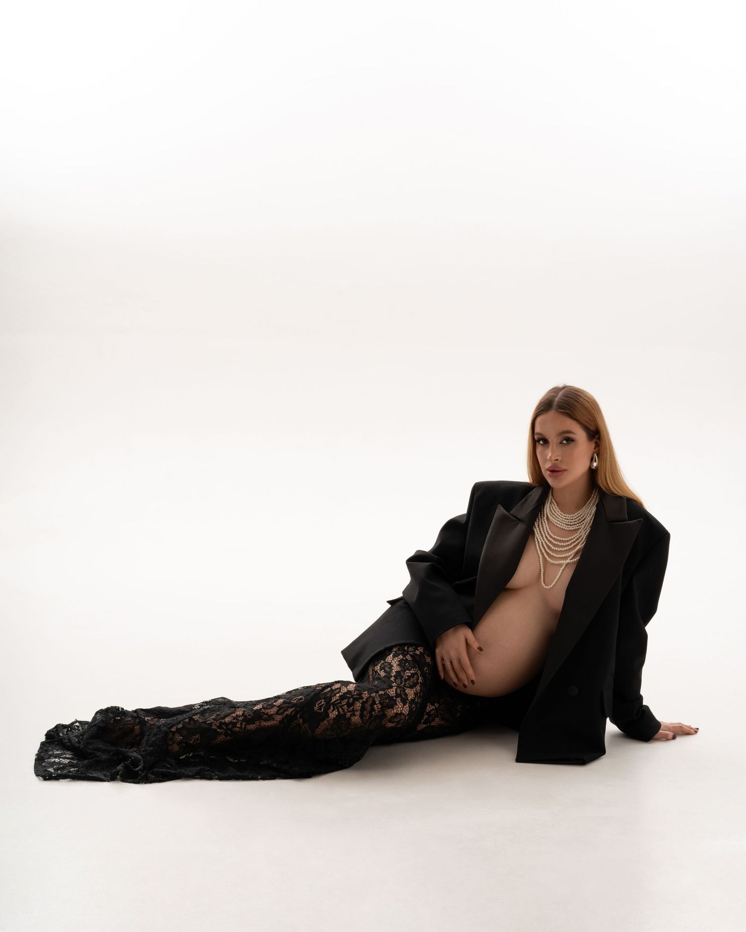 Maternity portrait of a stylish expecting mother in black jacket, black lace skirt and pearl necklace posing with confidence and grace.