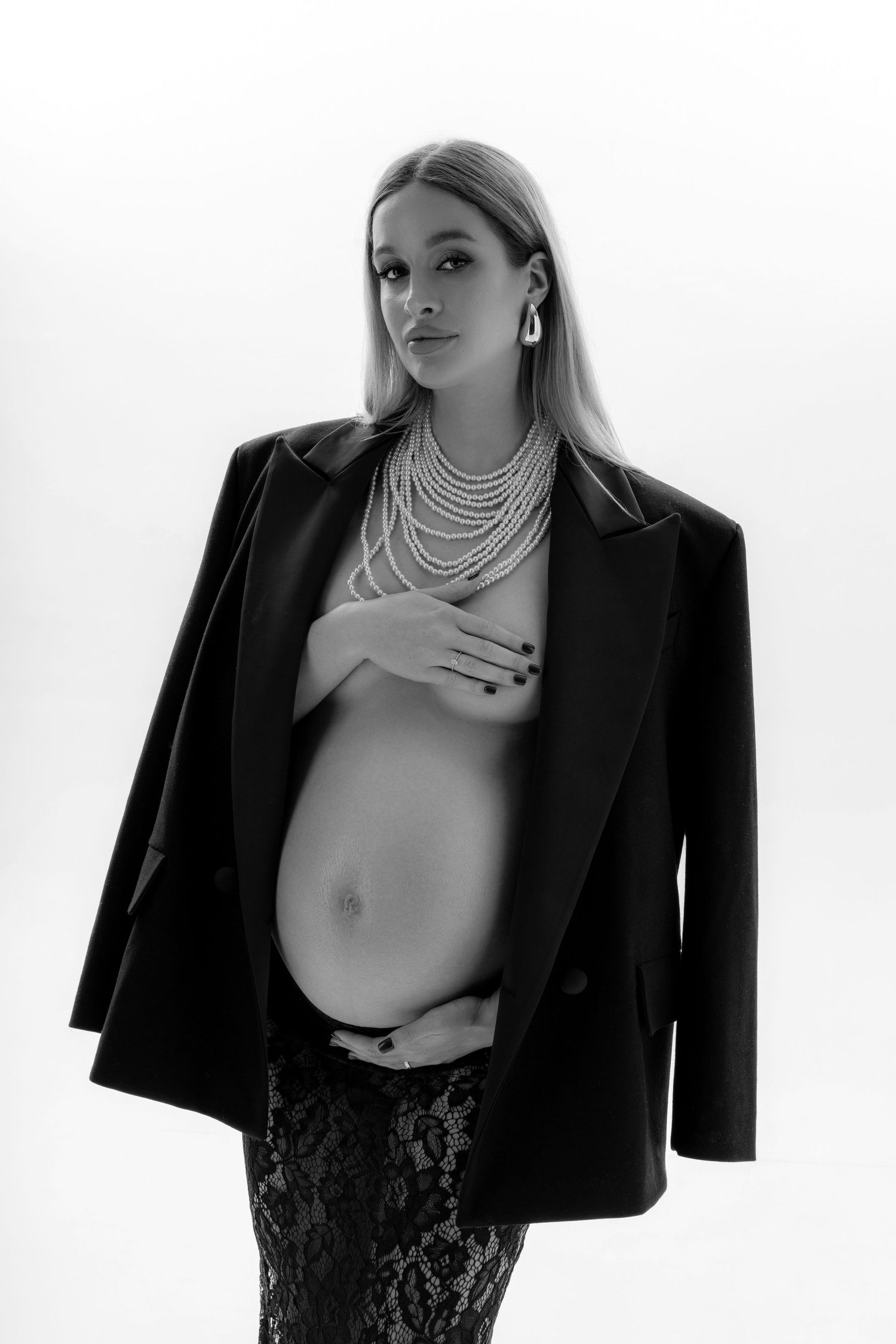 Artistic black and white maternity portrait capturing the pregnant women in black jacket, black lace skirt and pearl necklace.