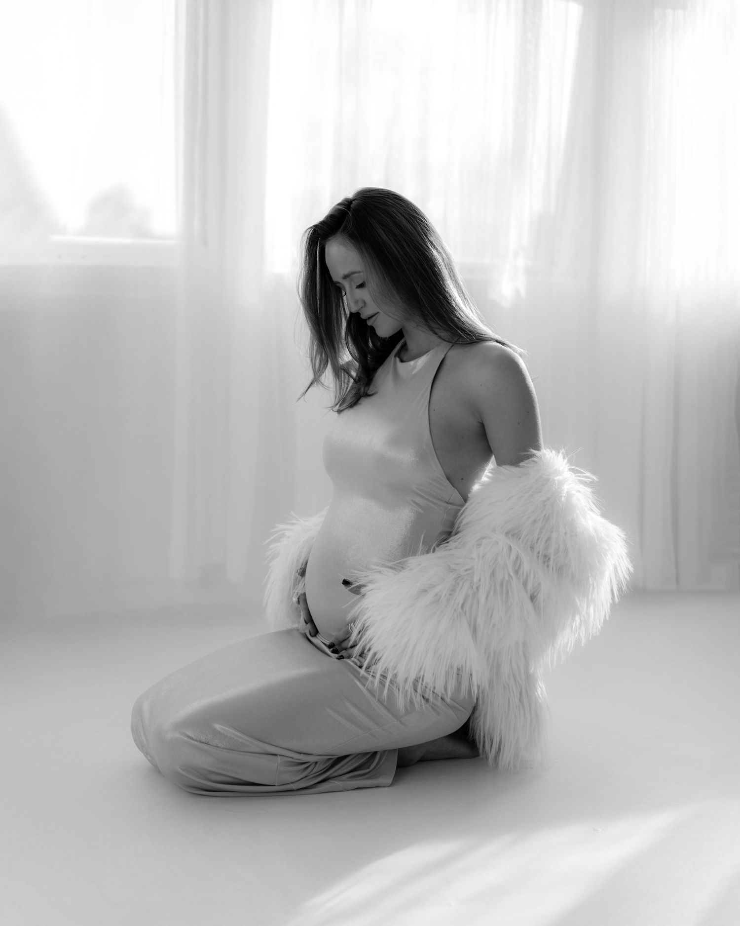 Black and white maternity portrait of an expecting mother in elegant dress and fur embracing her baby bump.