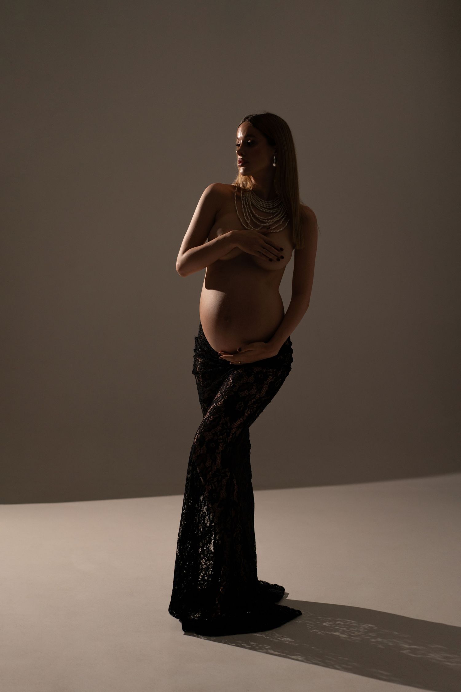 Maternity portrait of a stylish expecting topless mother in a long black lace skirt and pearl necklace posing with confidence and grace.