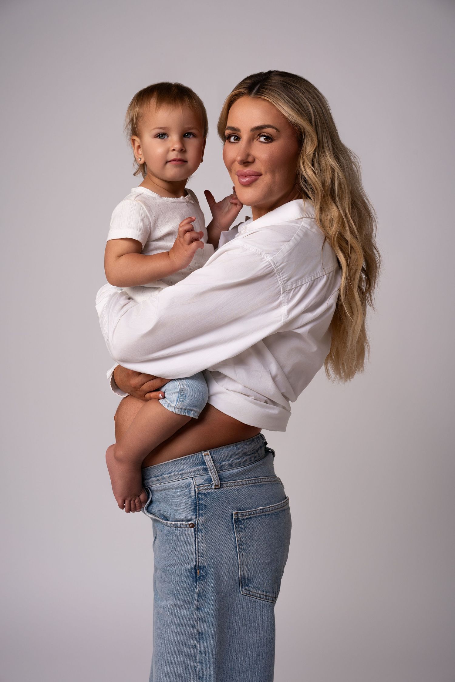 Pregnant mother and daughter in matching outfits posing during a maternity session — mom in white shirt and jeans holding her child.