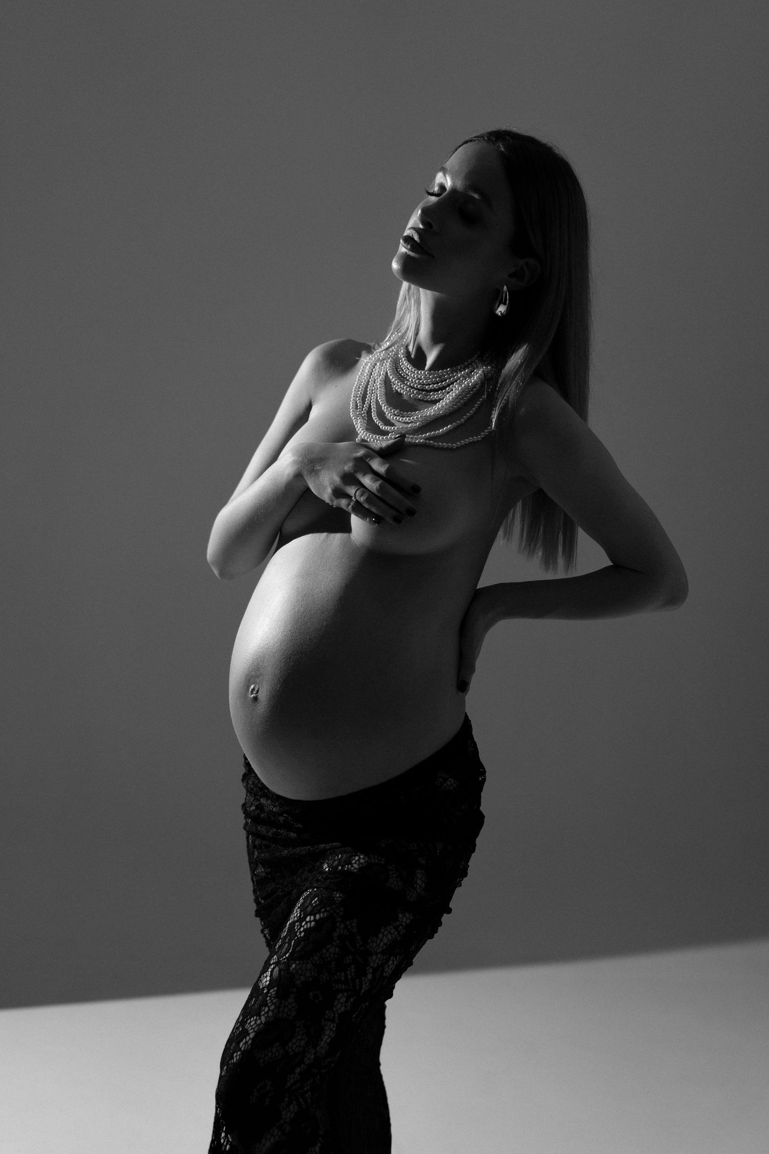 B&W maternity portrait of a stylish expecting topless mother in black lace skirt and pearl necklace posing with confidence and grace.
