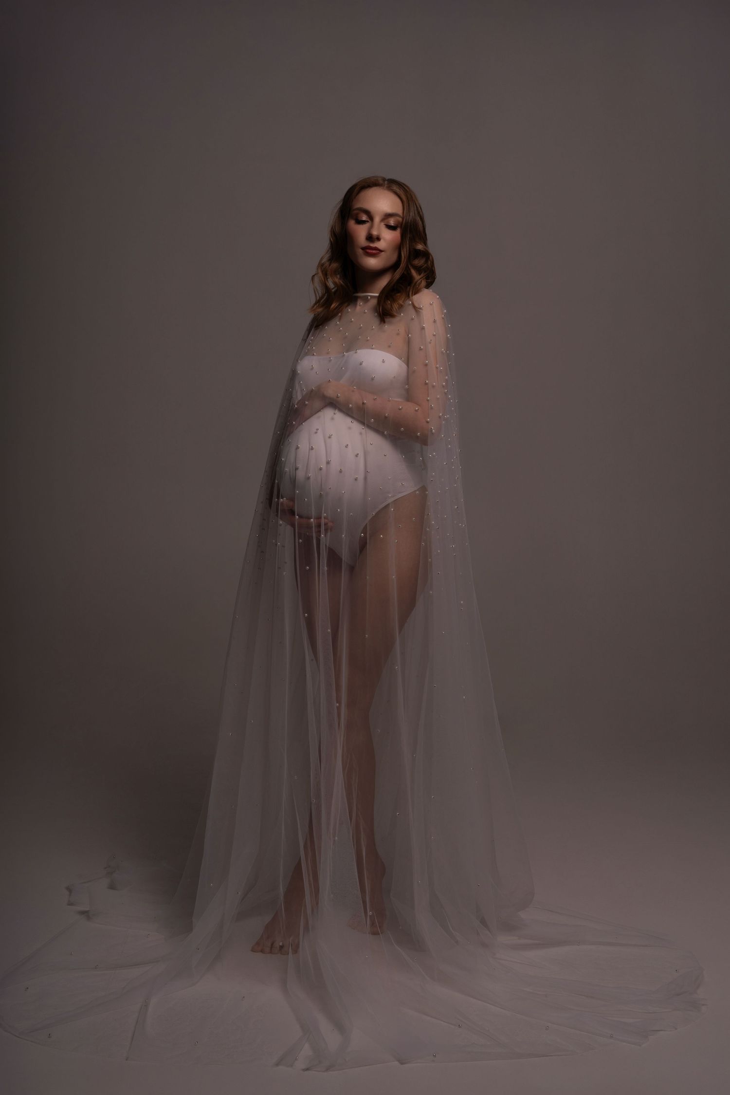 The pregnant women wearing white bodysuit and pearl cape posing in photo studio in Chicago.