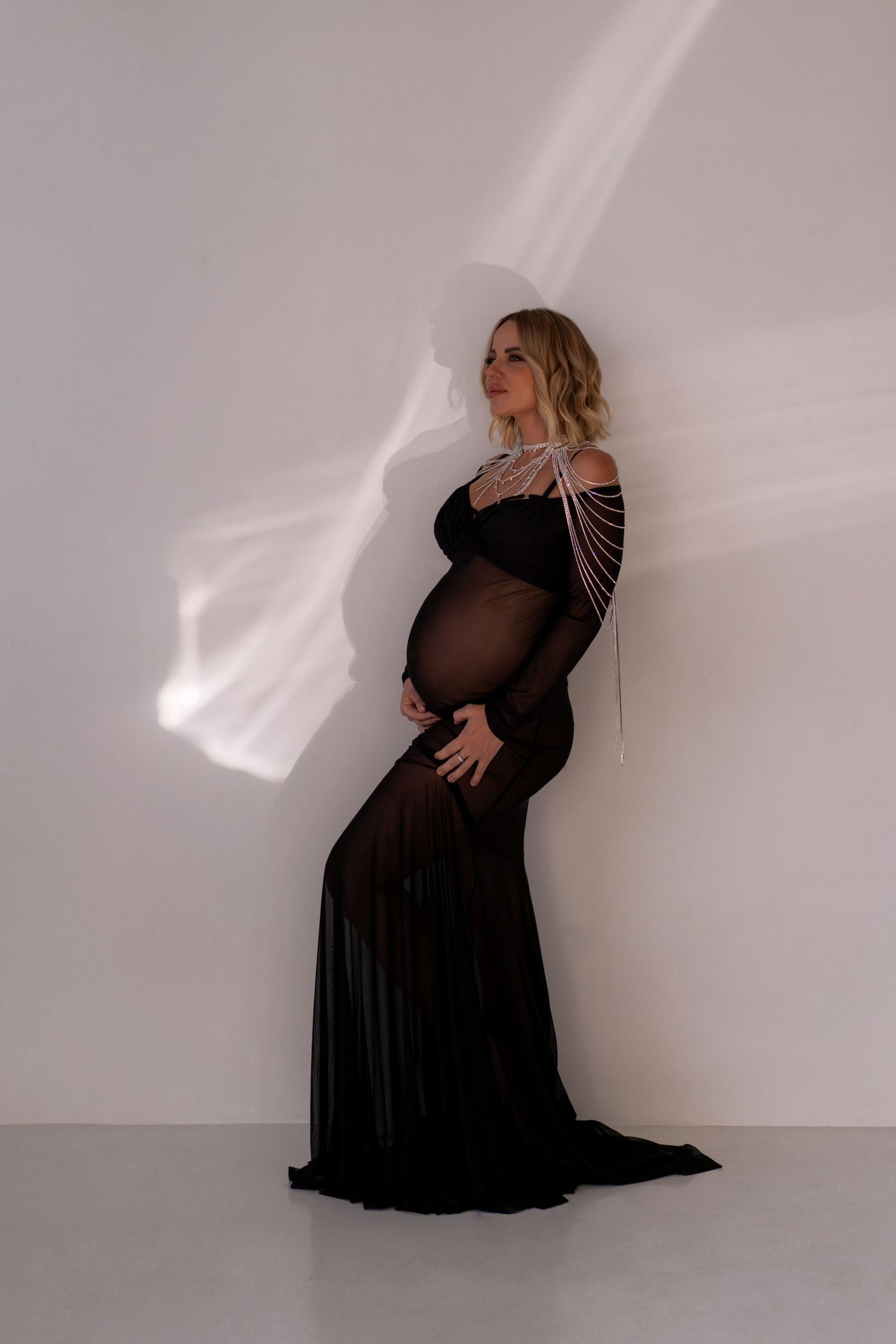 Glowing mother-to-be posing gracefully in a sheer black dress and crystal body chain against a neutral backdrop in Chicago photo studio.