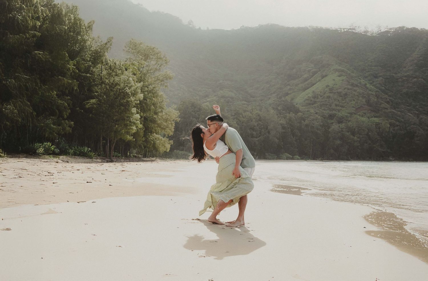 Couple sharing a romantic kiss on a tropical beach with lush green mountains in the background.