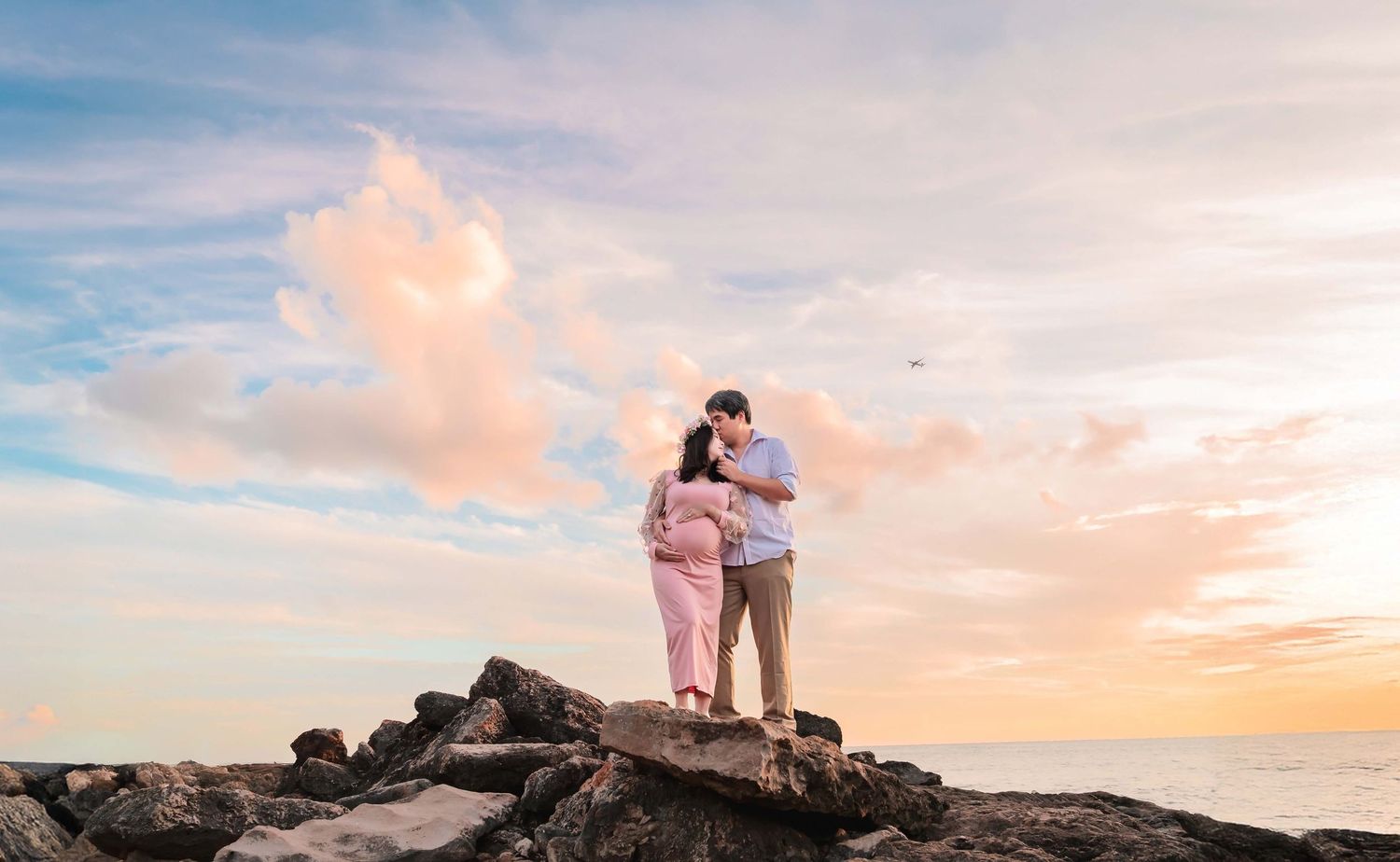 Couple embracing on rocky shore at sunset, woman in pink maternity dress, pastel sky with soft clouds in background.