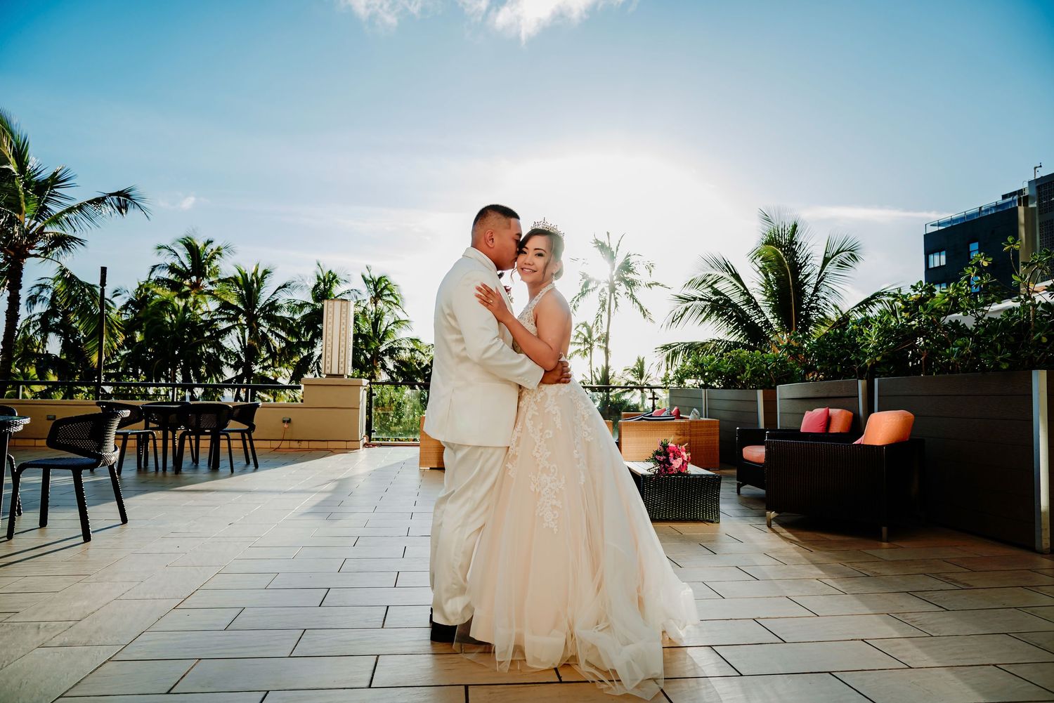 Bride and groom share romantic first dance on tropical rooftop terrace with palm trees and blue sky backdrop.