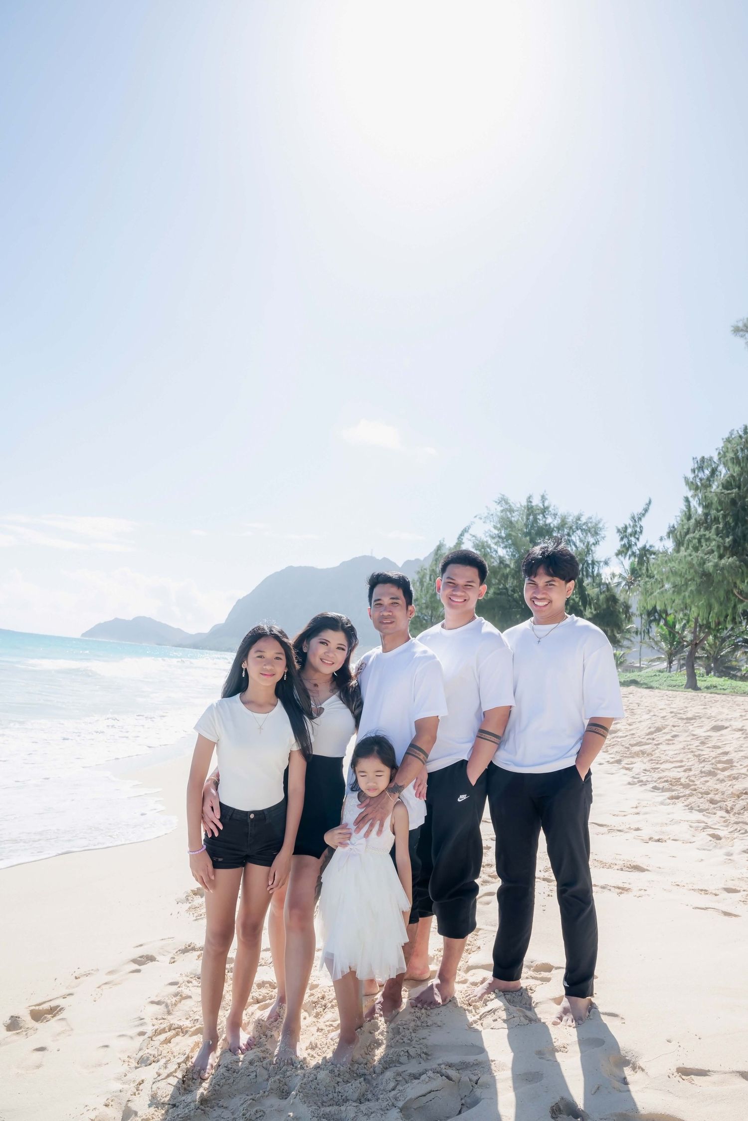 A joyful Asian family of six poses together on a sunny Hawaii beach with mountains and ocean in the background.