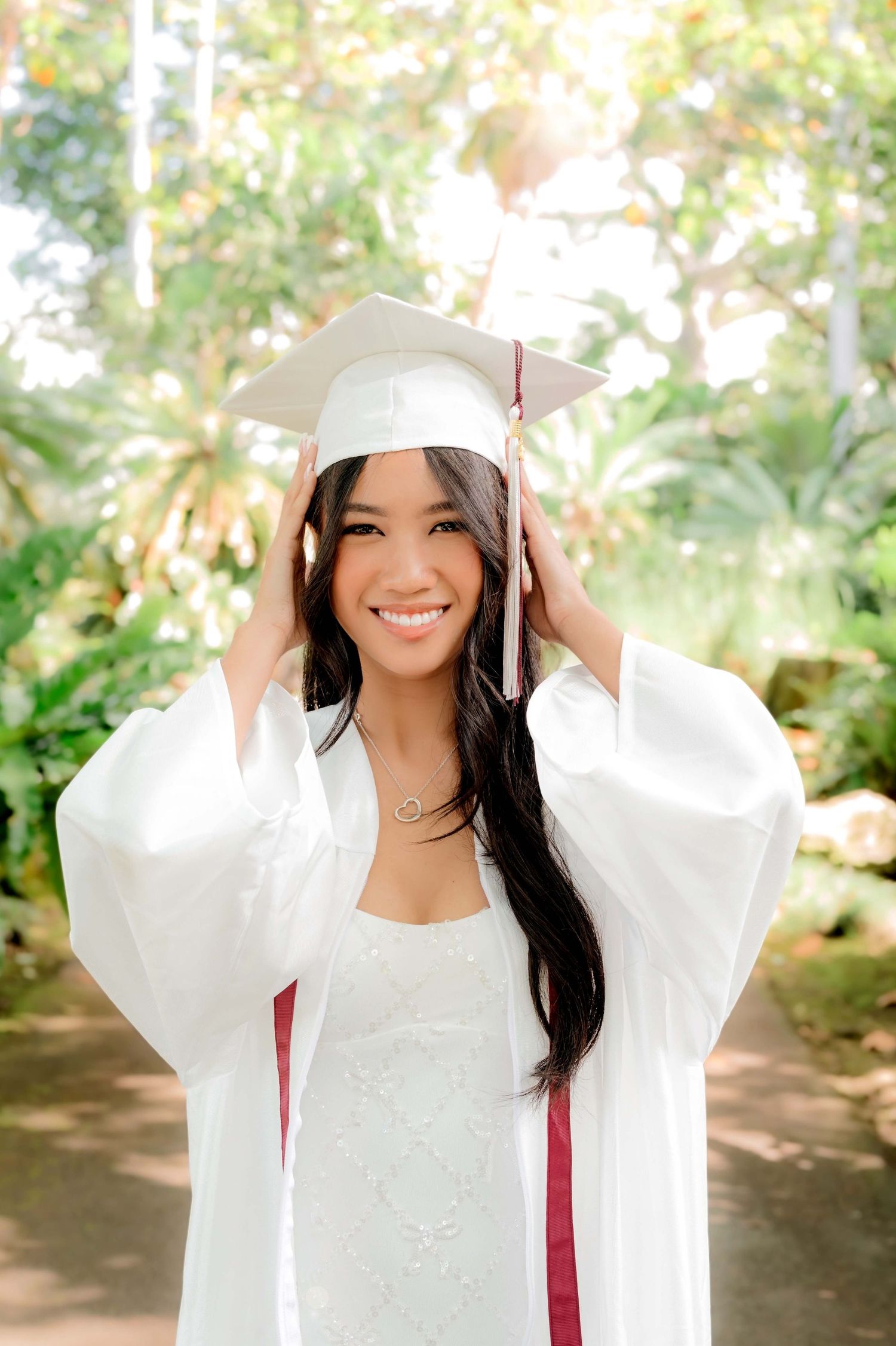 Young woman in white graduation cap and gown smiling outdoors with lush green trees in background.