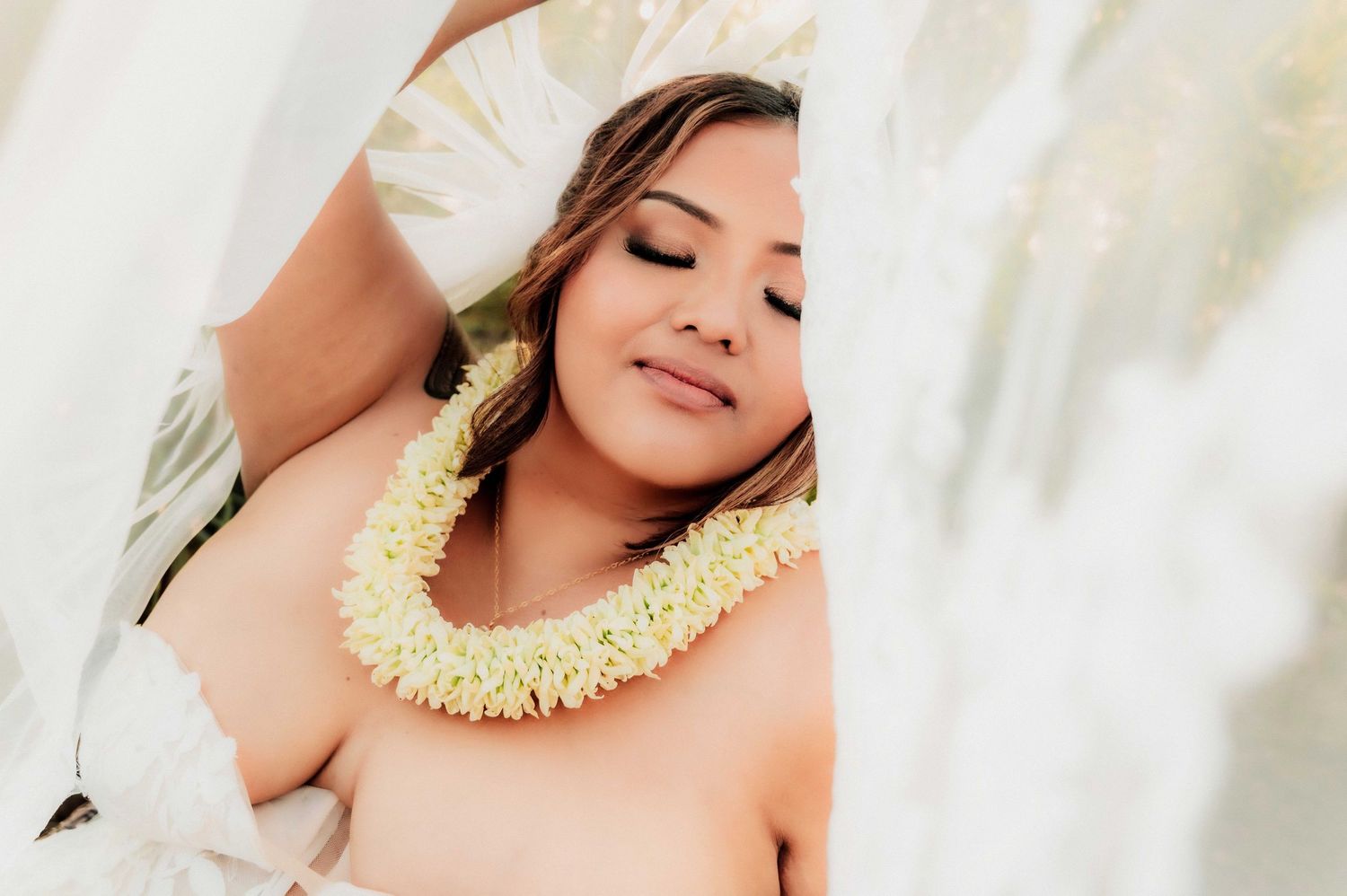 Woman wearing a white floral lei, posing gracefully with eyes closed among flowing white curtains.