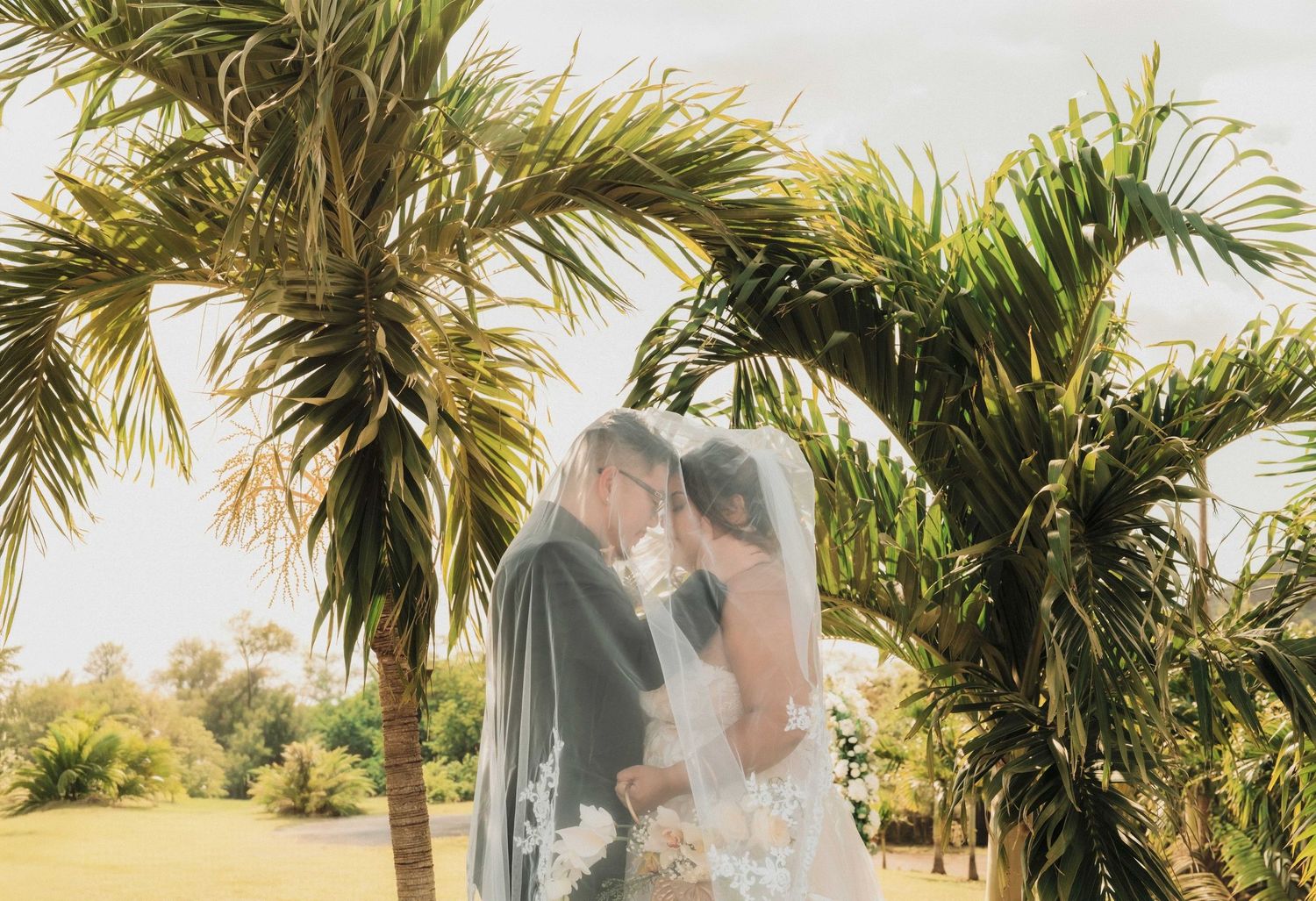 A bride and groom share a romantic kiss under lush tropical palm trees during an outdoor wedding ceremony.