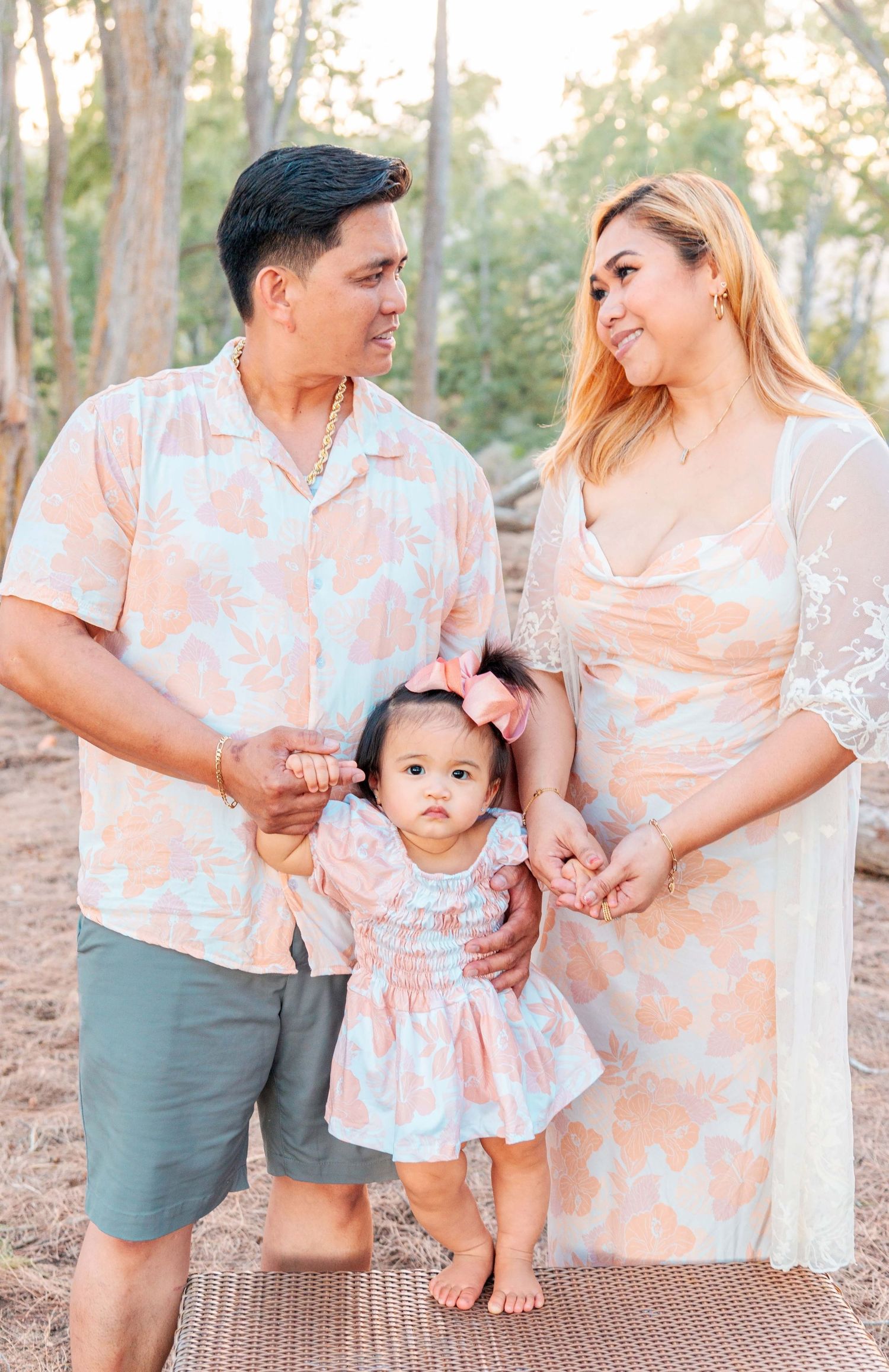 A family of three in matching peach floral outfits pose outdoors, parents holding hands with their toddler daughter.