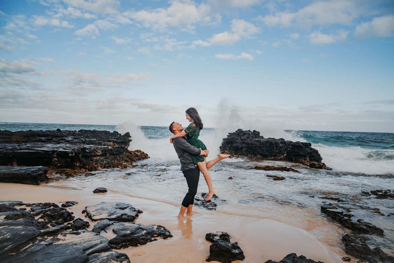 Couple embracing on rocky Hawaiian beach with crashing waves and dramatic ocean backdrop.
