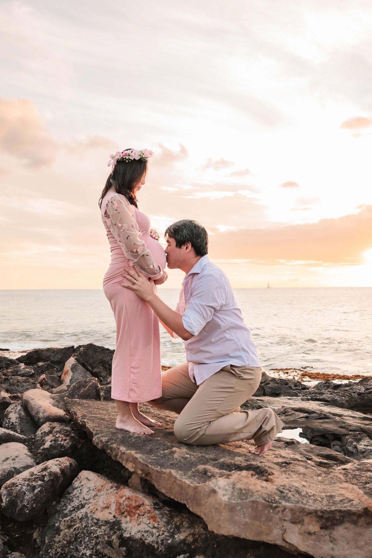 Couple sharing a tender maternity moment on rocky beach at sunset, woman in pink dress with floral crown, man kneeling.