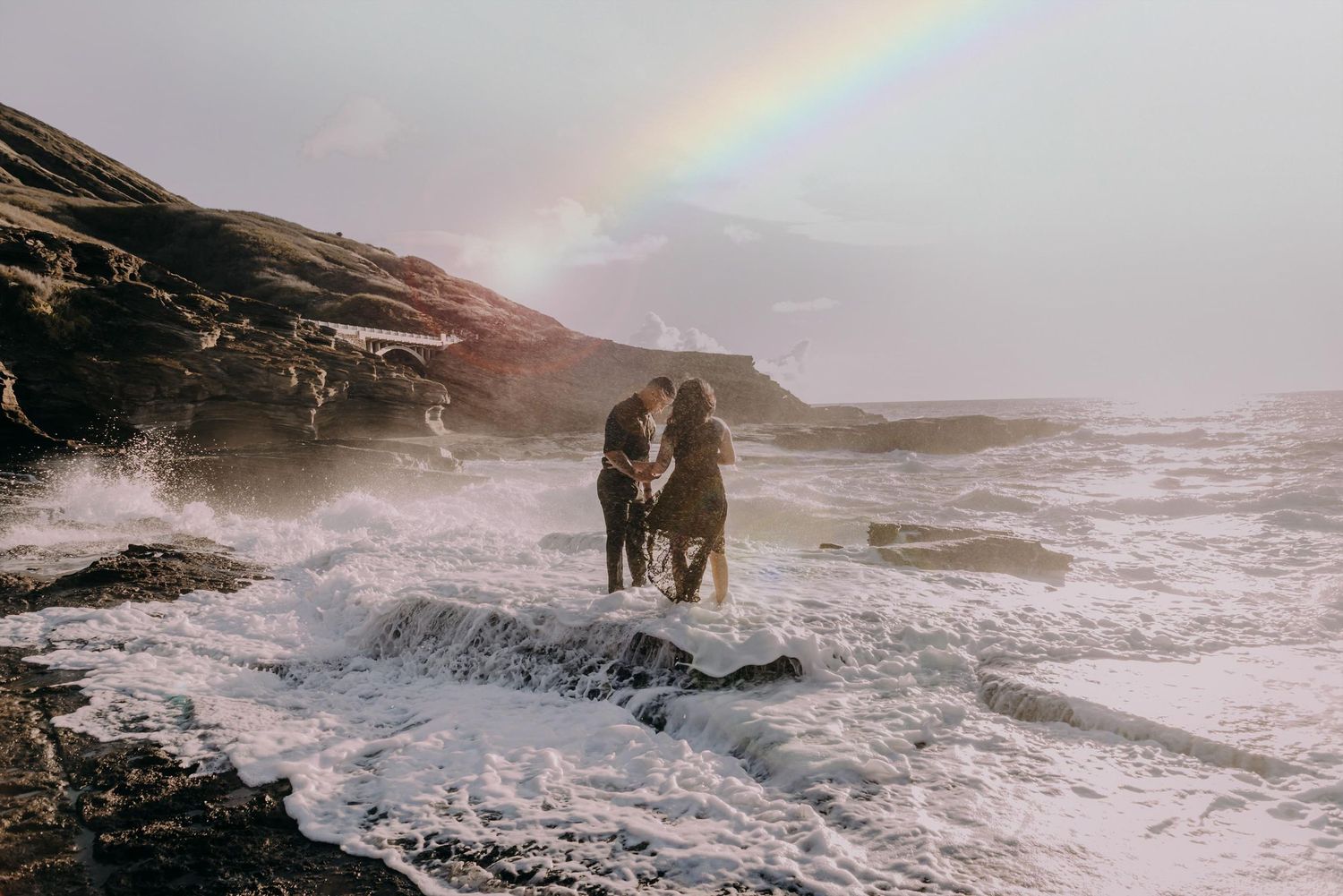 A beautiful rainbow arches over a dramatic rocky coastline as waves crash against the shore during sunset.