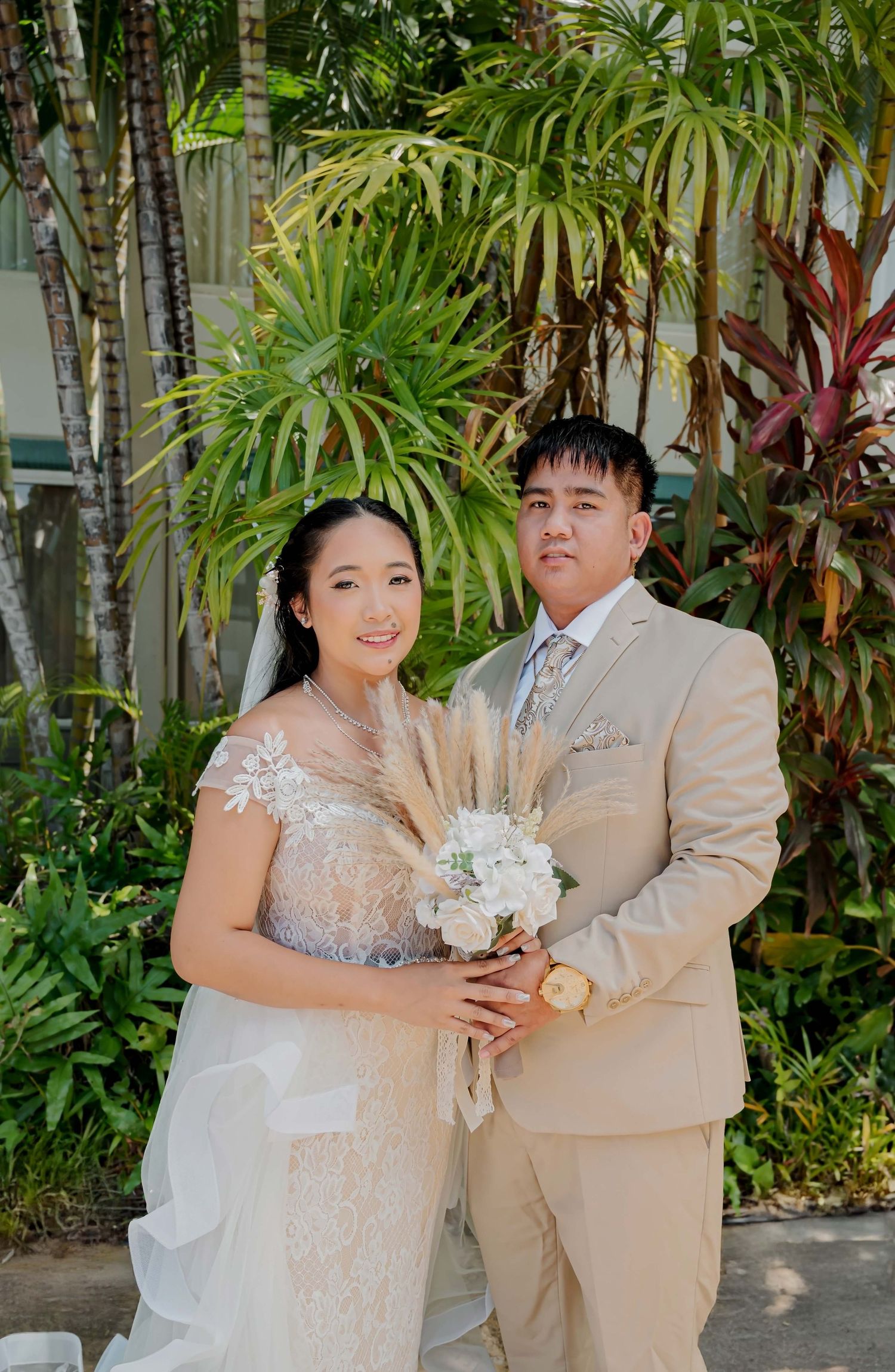 Couple poses for wedding photos in a tropical garden setting wearing beige and cream formal attire.