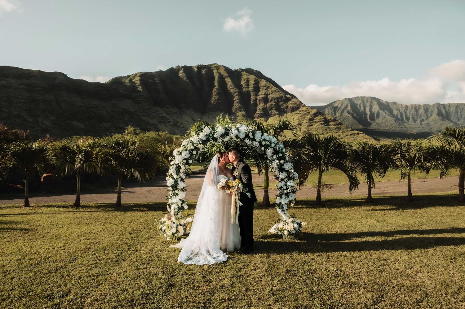 Couple exchanging vows under floral arch with lush green mountains backdrop at outdoor Hawaii wedding ceremony.