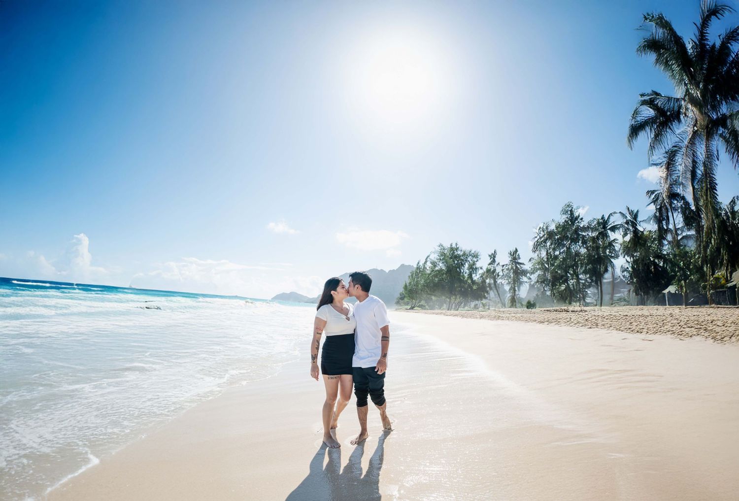 Couple kissing on a sunny tropical beach with white sand, turquoise waves, and palm trees in the background.
