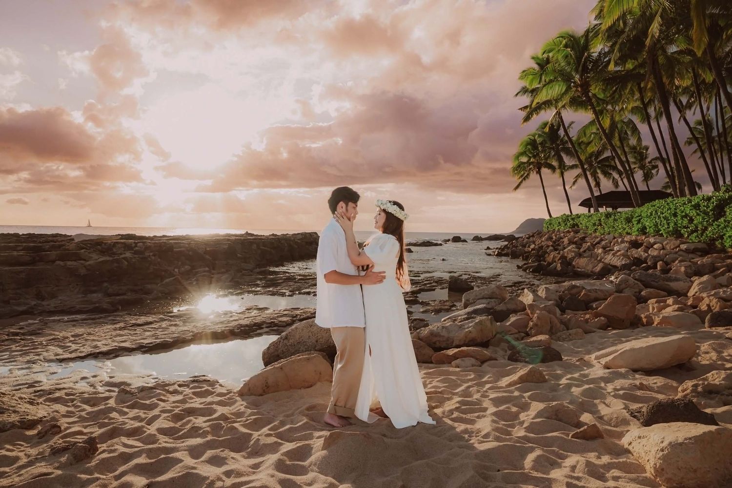 Couple in white attire embracing on a rocky Hawaiian beach at sunset with palm trees and dramatic sky.