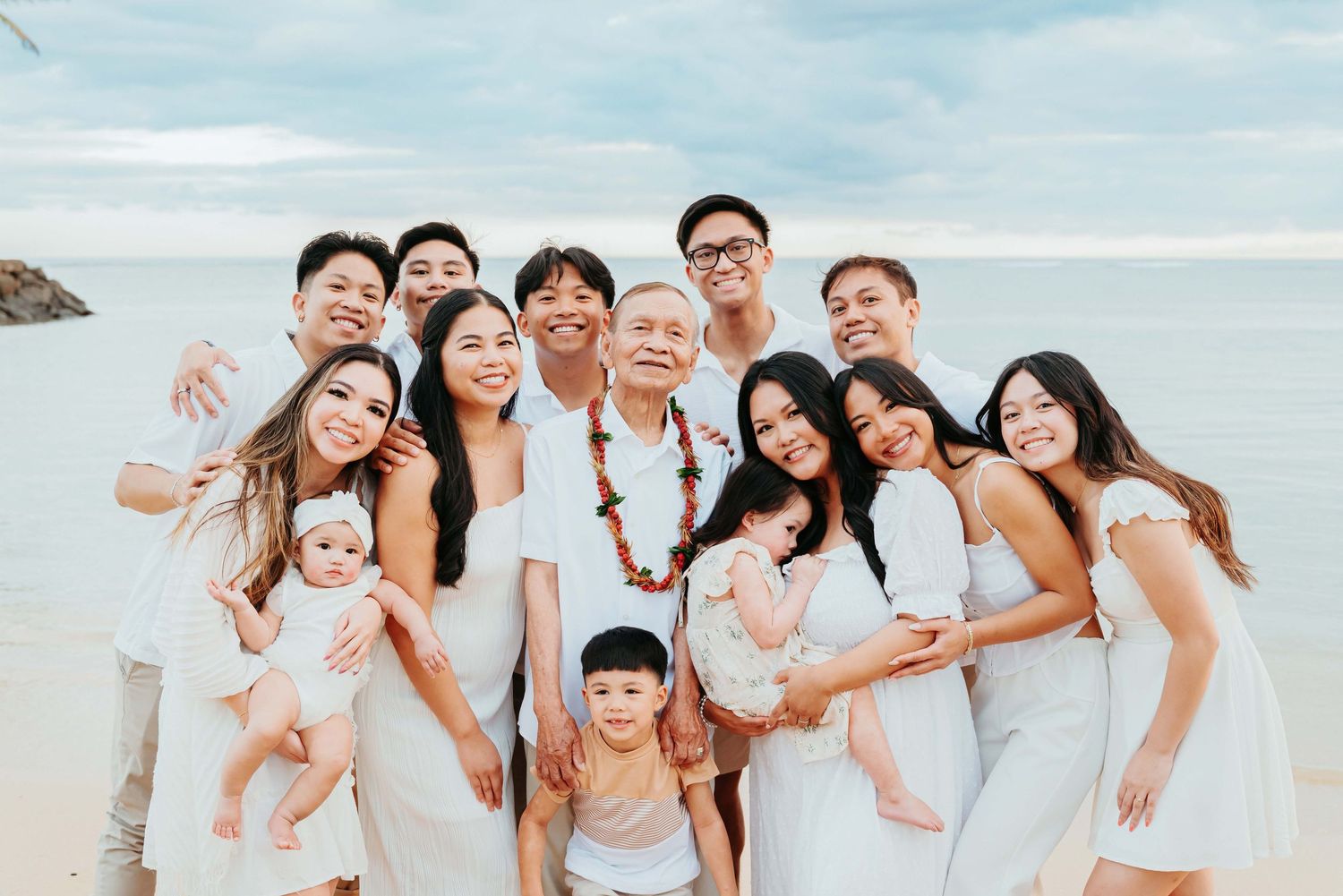 Large Asian family in white outfits posing together on a tropical beach with turquoise water in the background.