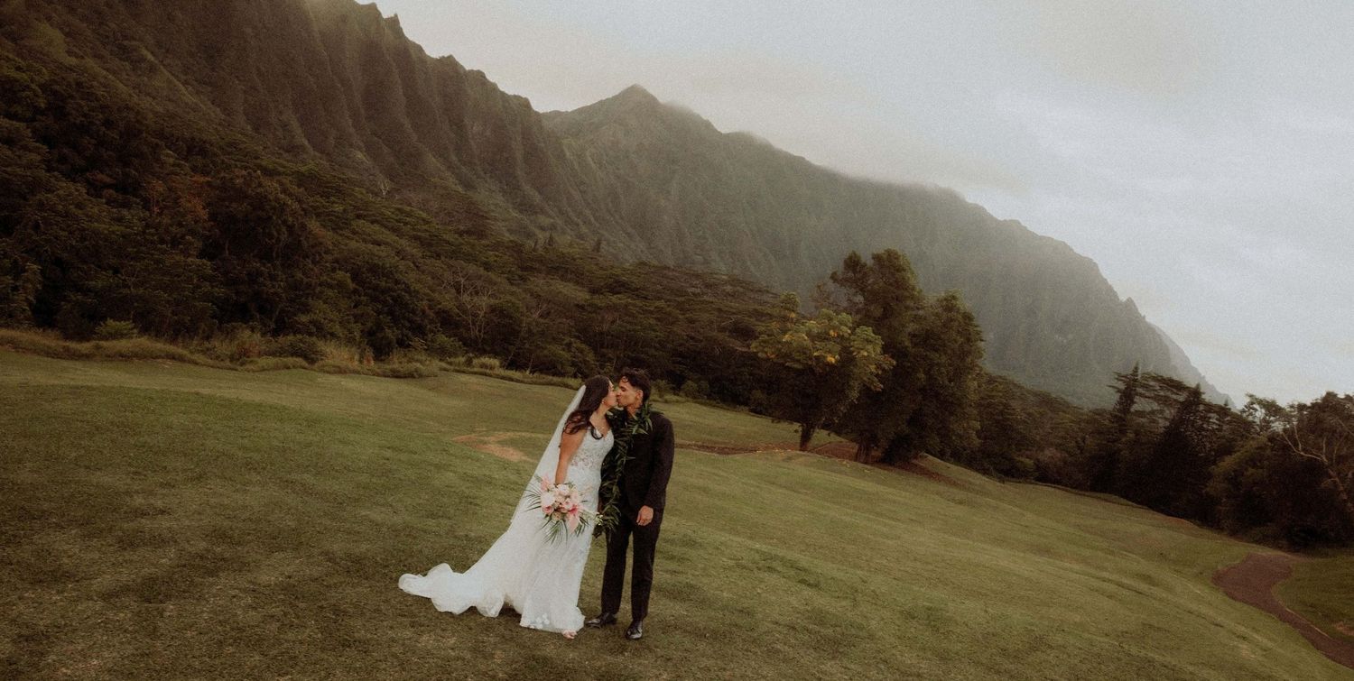 Couple in wedding attire standing on lush green lawn with dramatic misty mountain cliffs in Hawaii backdrop.