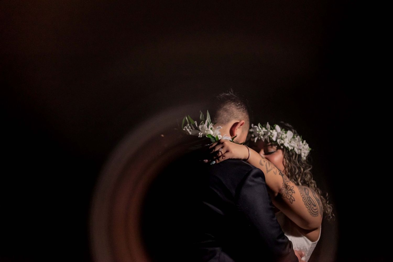 Bride and groom share a romantic kiss under dramatic dark lighting with a floral crown at their wedding reception.