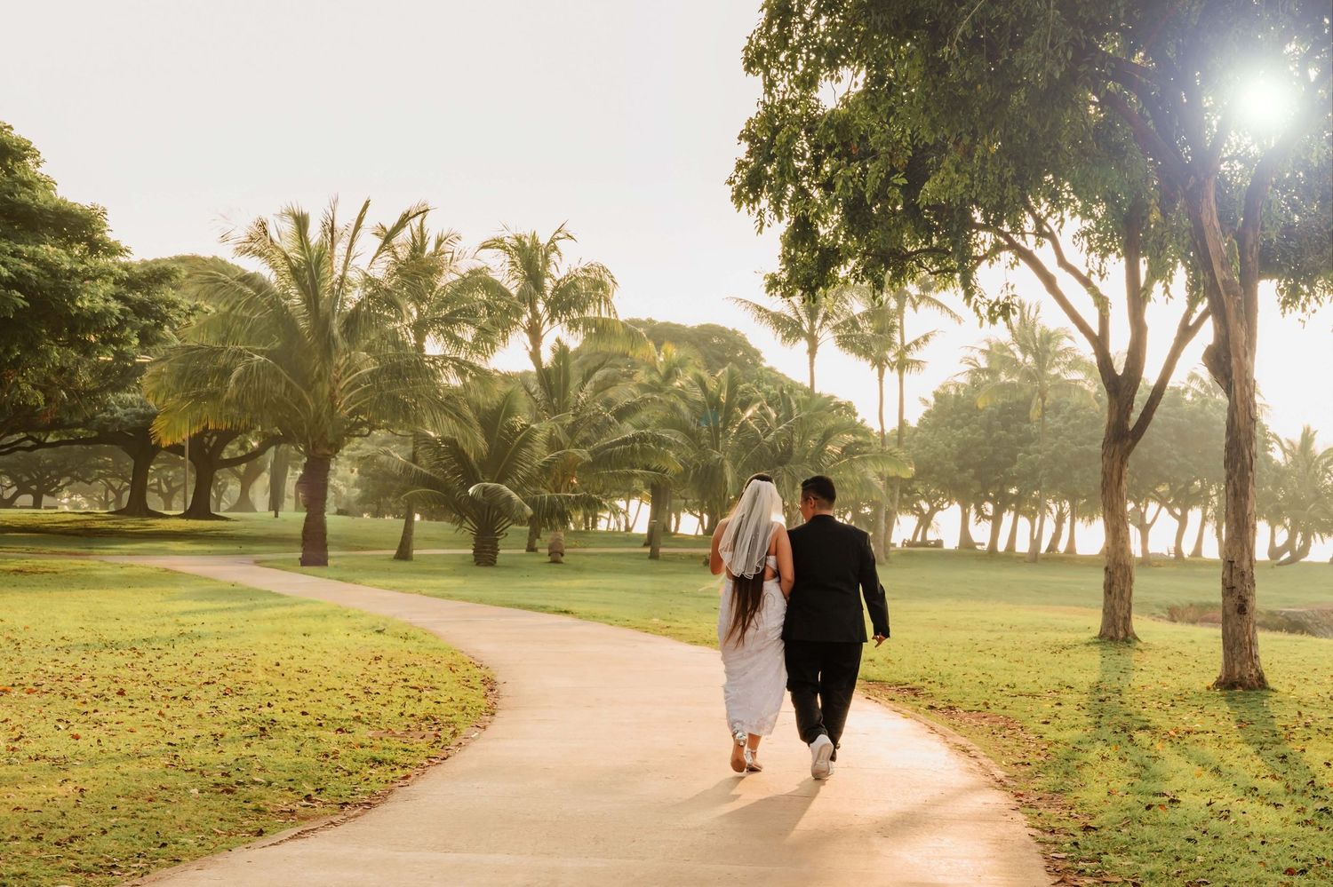 Bride and groom walking hand in hand along a tropical park path surrounded by palm trees at sunset.