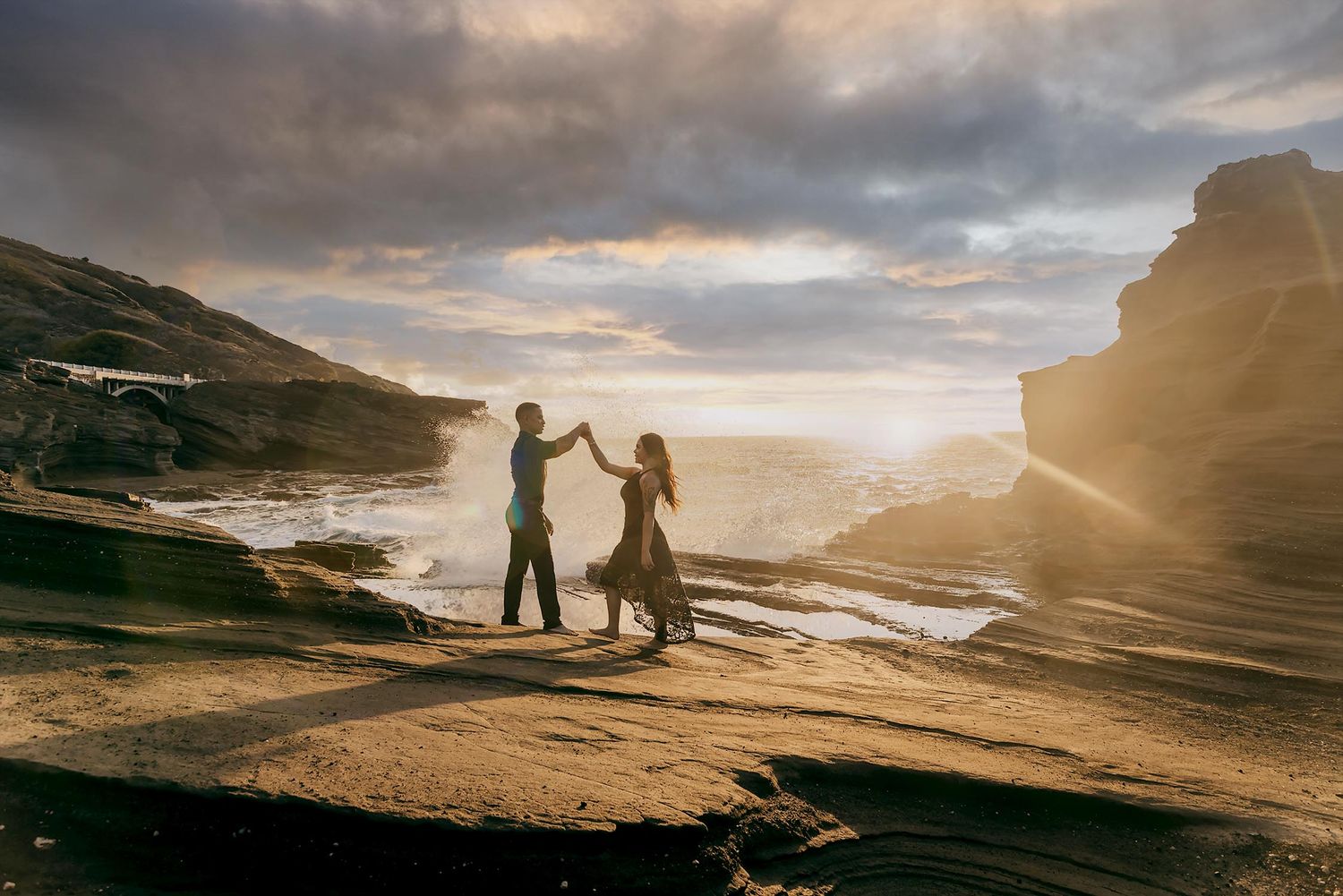 Couple dancing on rocky coastal cliffs at golden sunset with dramatic ocean waves and glowing sky backdrop.