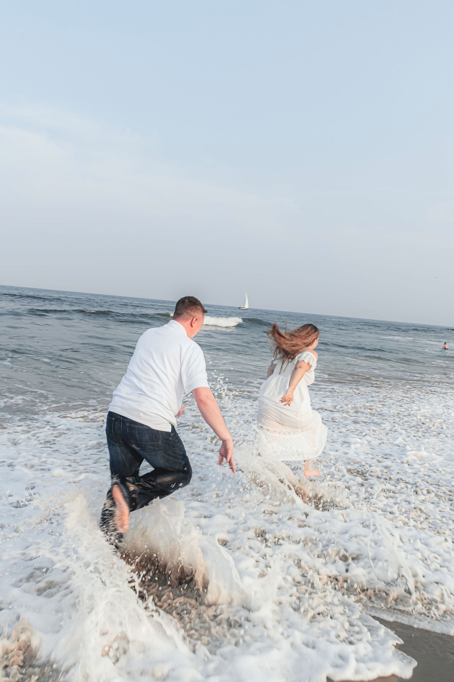A couple in formal attire gets splashed by an ocean wave while playing at the beach during a photoshoot.