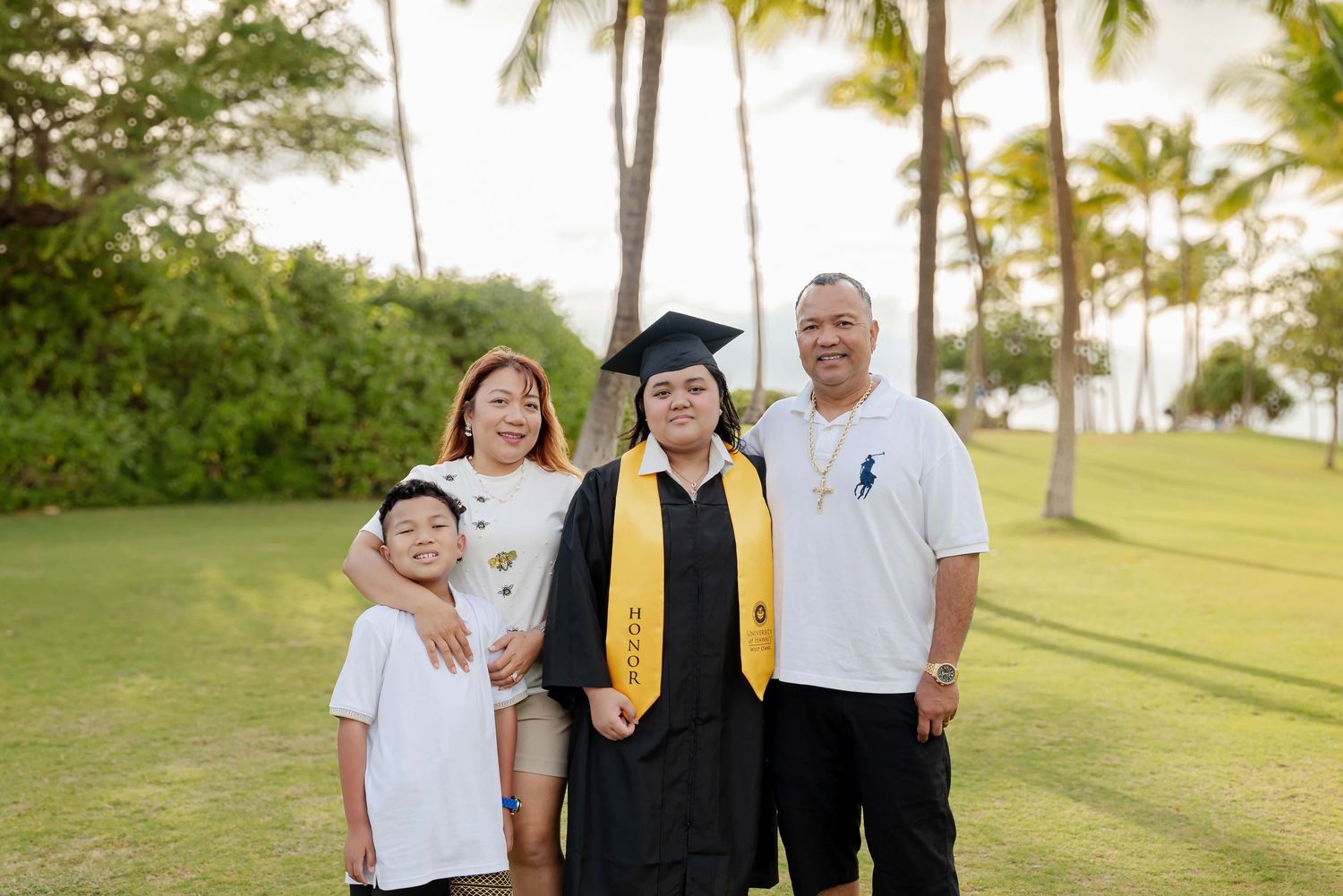 Family of four poses outdoors on green lawn with palm trees, celebrating womans graduation in cap and gown with yellow sash.