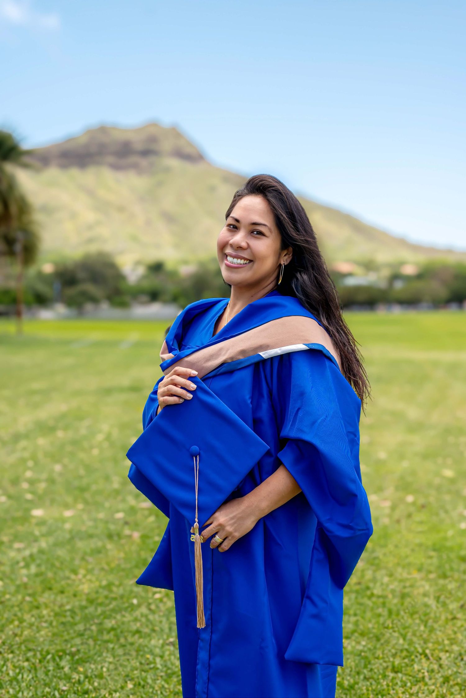 Smiling woman in blue graduation gown and cap poses outdoors on green field with mountain backdrop.