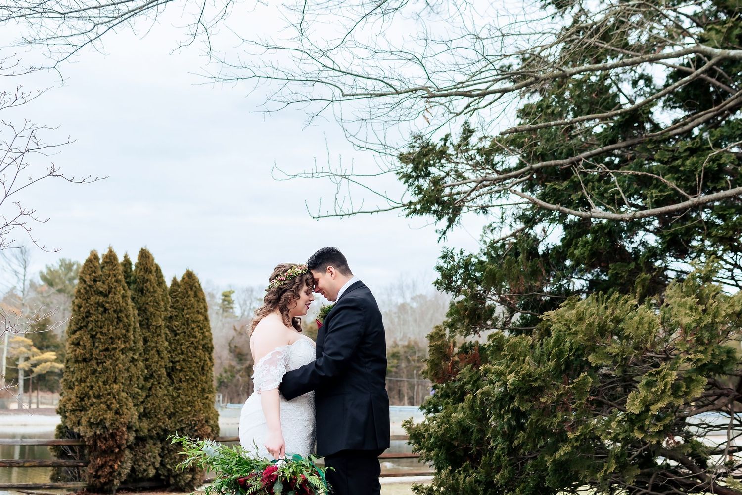 A couple in winter wedding attire embrace outdoors near evergreen trees and snow dusted branches.