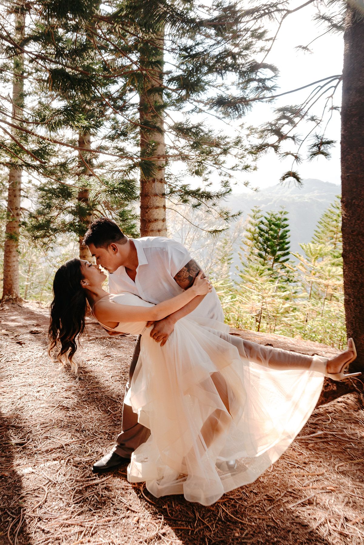A couple in wedding attire share a romantic dip pose on a forest trail with sunlit evergreen trees in the background.