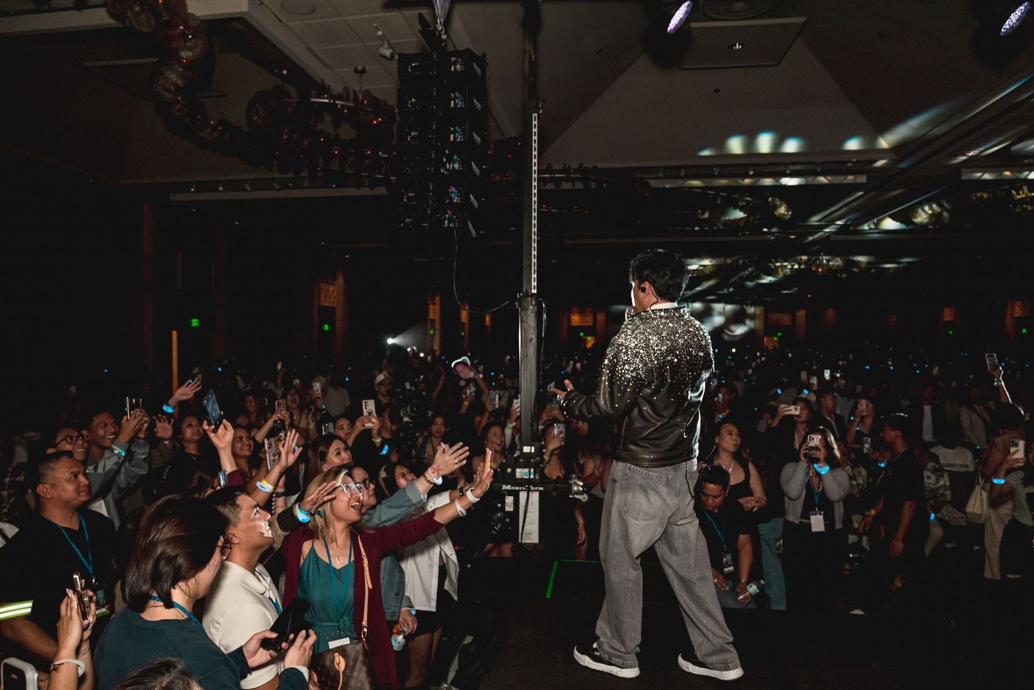 Performer on stage with back to camera engaging energetic crowd at indoor concert venue with dramatic lighting.