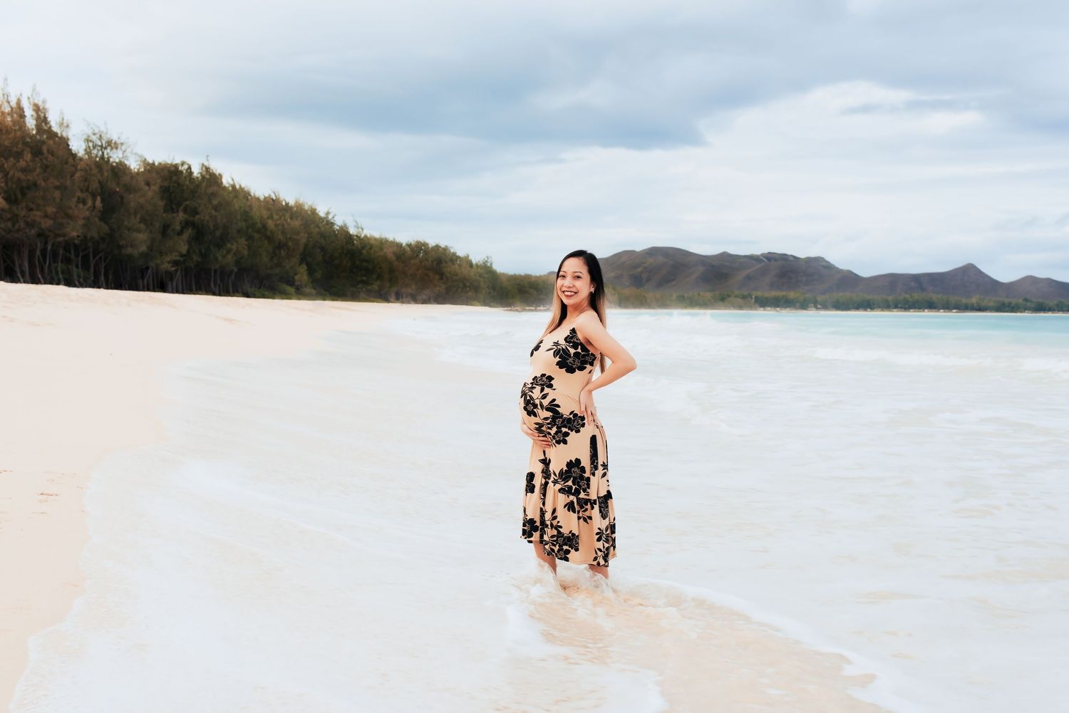 Pregnant woman in floral dress standing on a tropical white sand beach with turquoise water and green mountains.