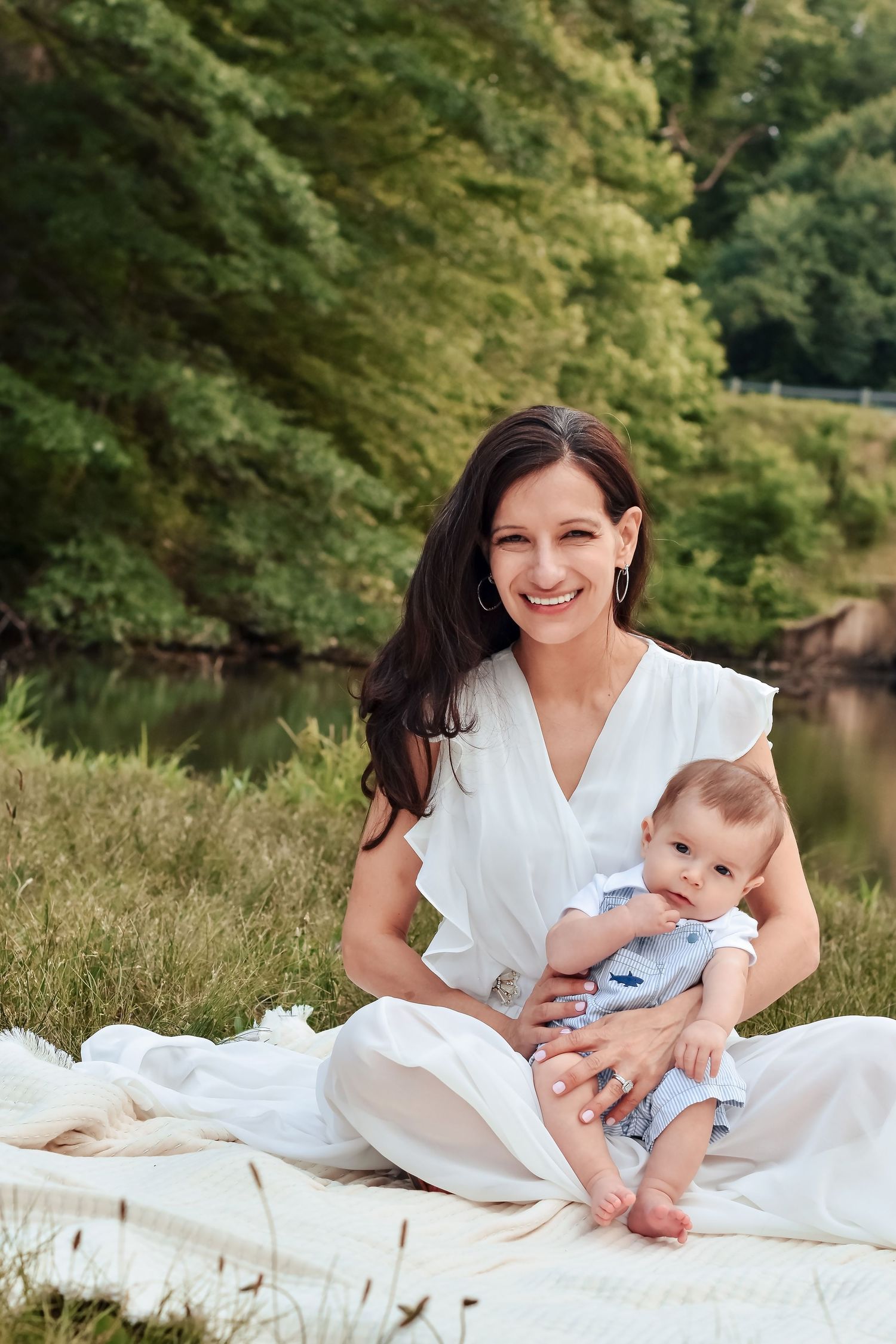 Smiling brunette woman in white dress sitting on blanket outdoors holding a baby in blue outfit near a river.