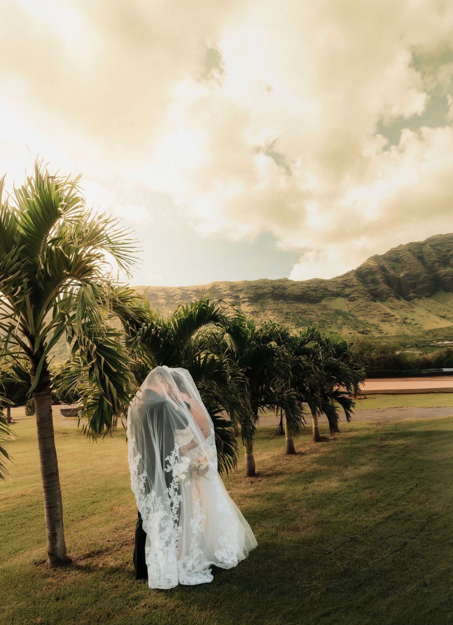 Bride in lace wedding gown and veil stands among palm trees with lush green mountains backdrop in Hawaii.