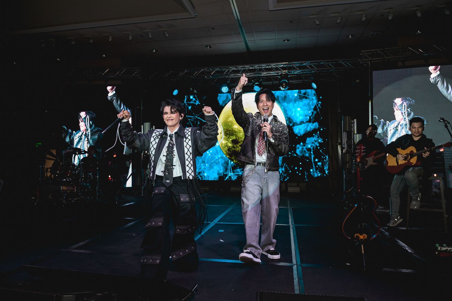 Two performers raise their hands on stage during a live concert with band members and blue LED backdrop.