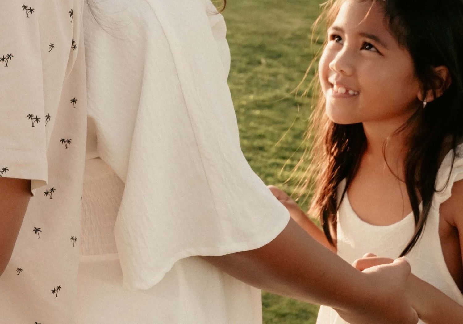 A young girl in a white dress smiles up at an adult wearing a floral skirt outdoors on green grass.