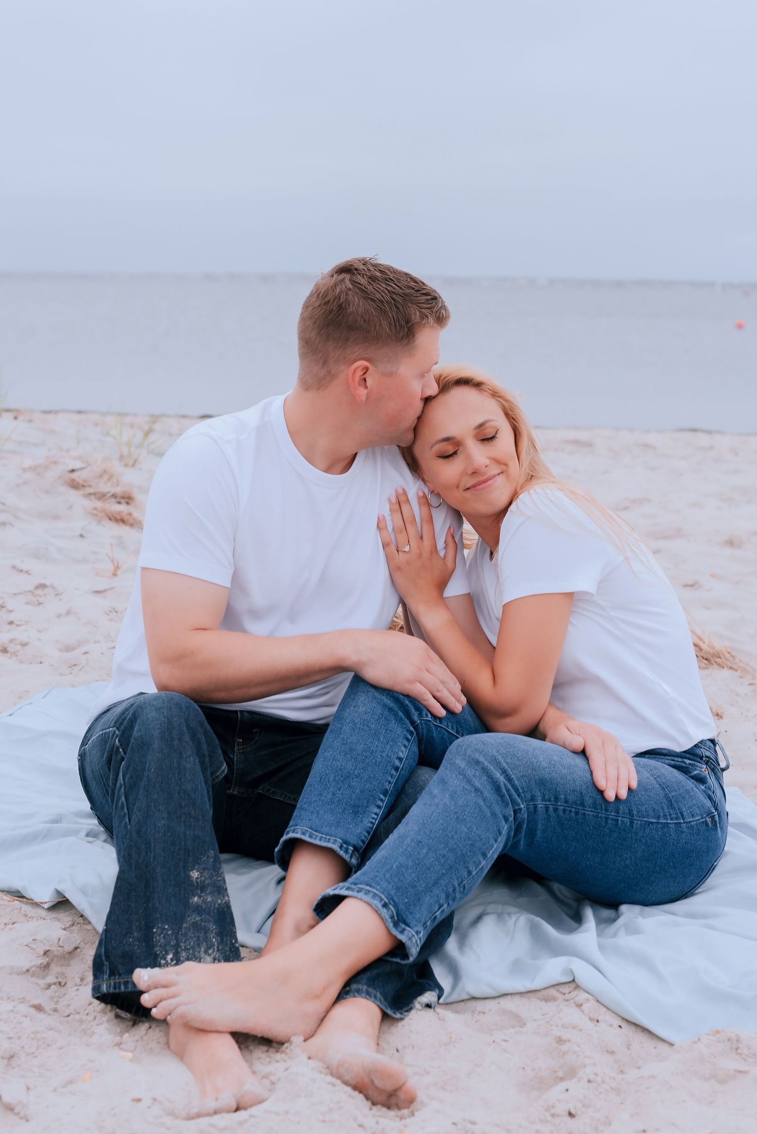 Couple sitting on sandy beach blanket, man kissing woman's temple, both wearing white tees and jeans, ocean backdrop.
