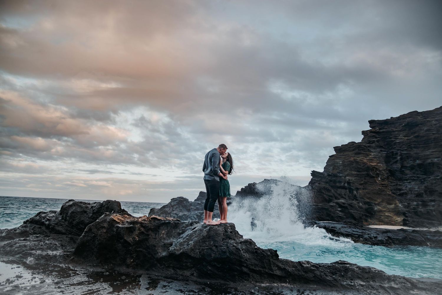 Couple embracing on rocky volcanic coastline with dramatic ocean waves crashing and cloudy sunset sky in Hawaii.