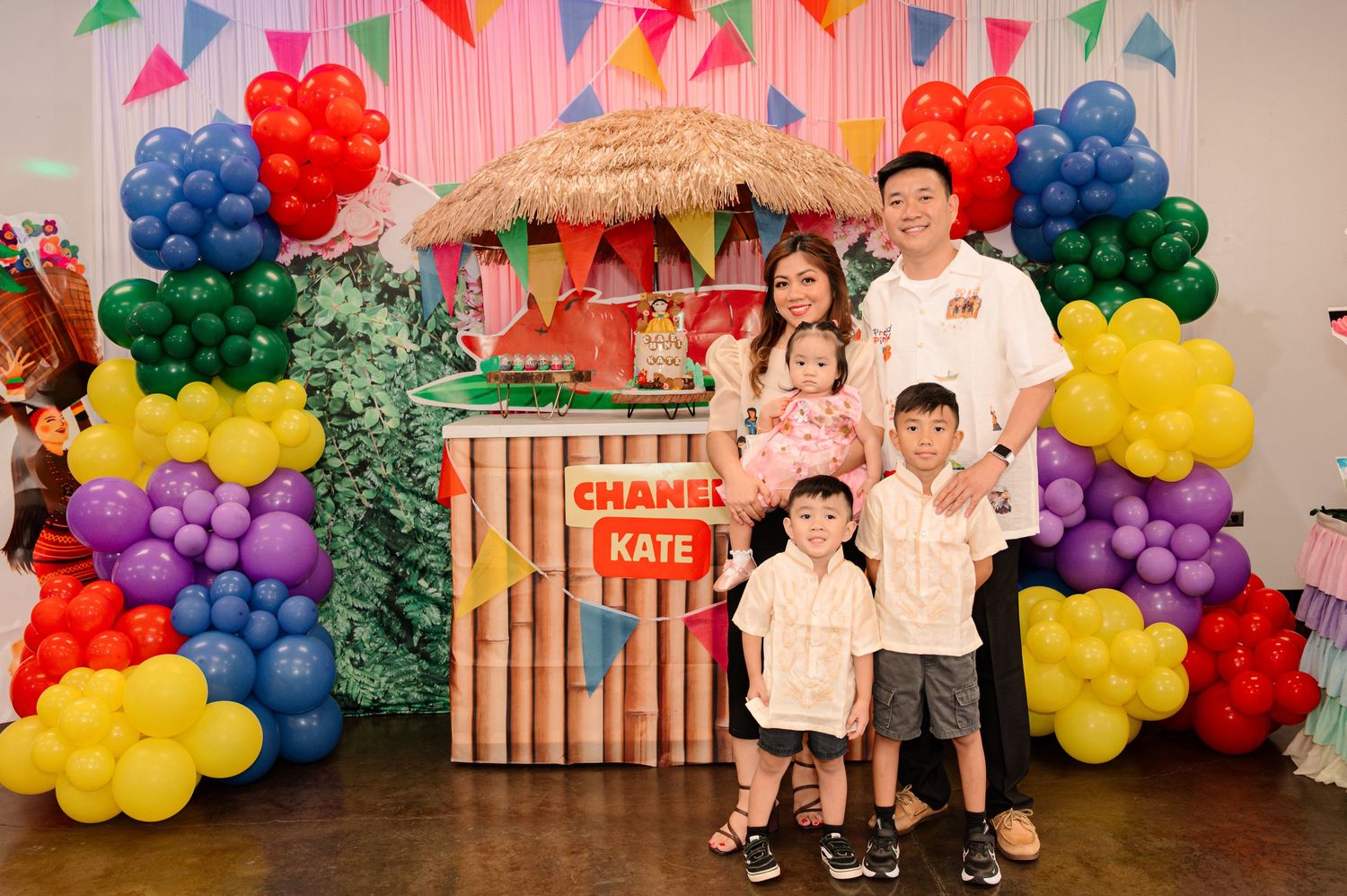 Family posing at a colorful birthday party with balloon arch, tropical decor, and a sign reading Chans Kate.