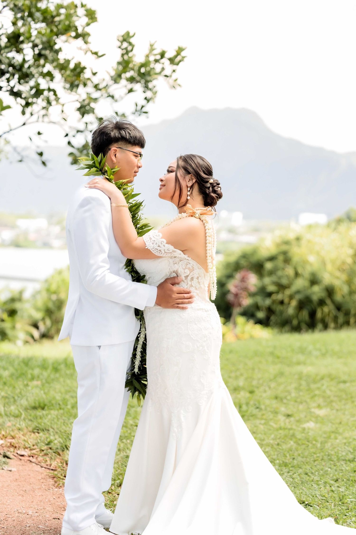Bride and groom in white attire embrace outdoors with lush greenery and mountain backdrop at Hawaiian wedding.