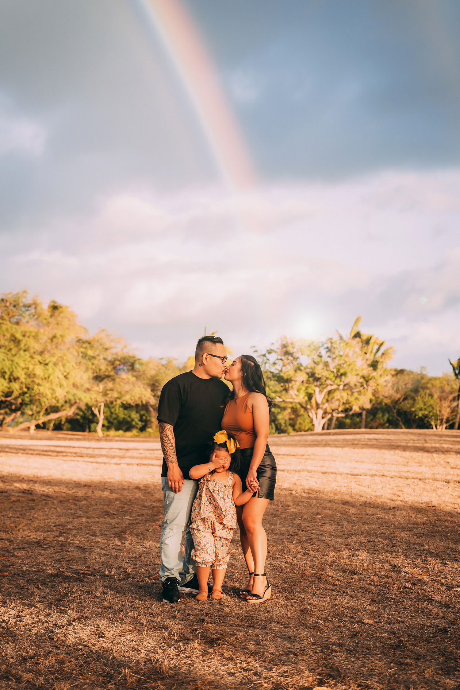 A couple shares a kiss with their child beneath a vibrant rainbow in a golden-lit outdoor setting.