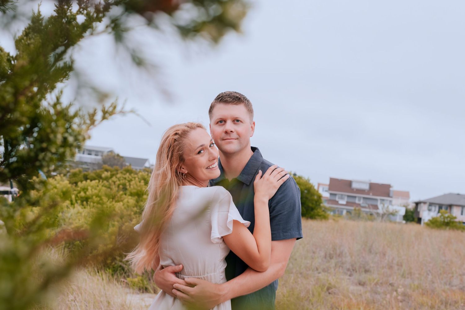 Couple embracing outdoors in a coastal field, woman in white dress, man in green polo shirt, with houses in background.